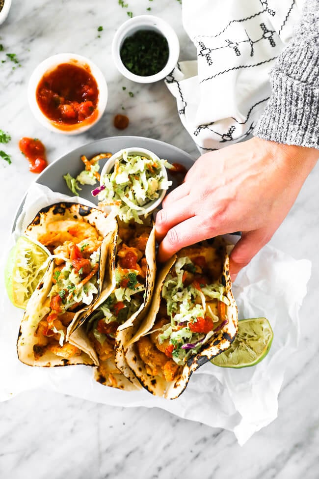 Overhead vertical image of hand coming in to pick up one taco from the plate of fish tacos. 