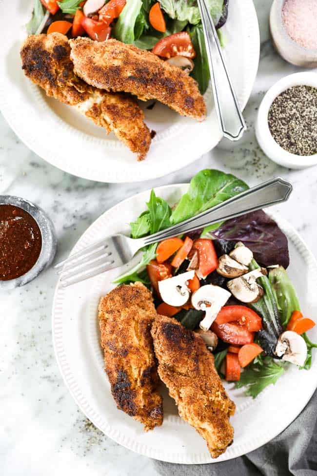 Paleo fried chicken served on two plates with side salads made with mixed greens, chopped carrots, tomatoes and mushrooms. BBQ dipping sauce on the side. 