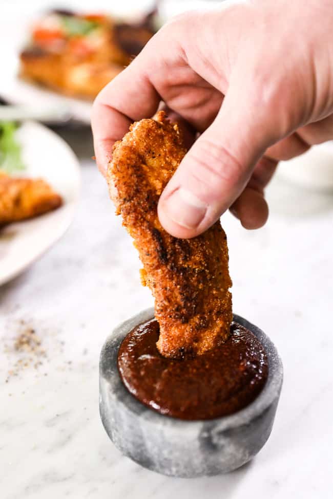 A piece of paleo fried chicken being dipped into ramekin of BBQ sauce. 