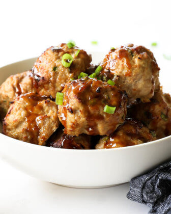 Angled image of a teriyaki turkey meatballs piled in a bowl with sticky teriyaki sauce drizzled on top. Garnished with chopped green onion.