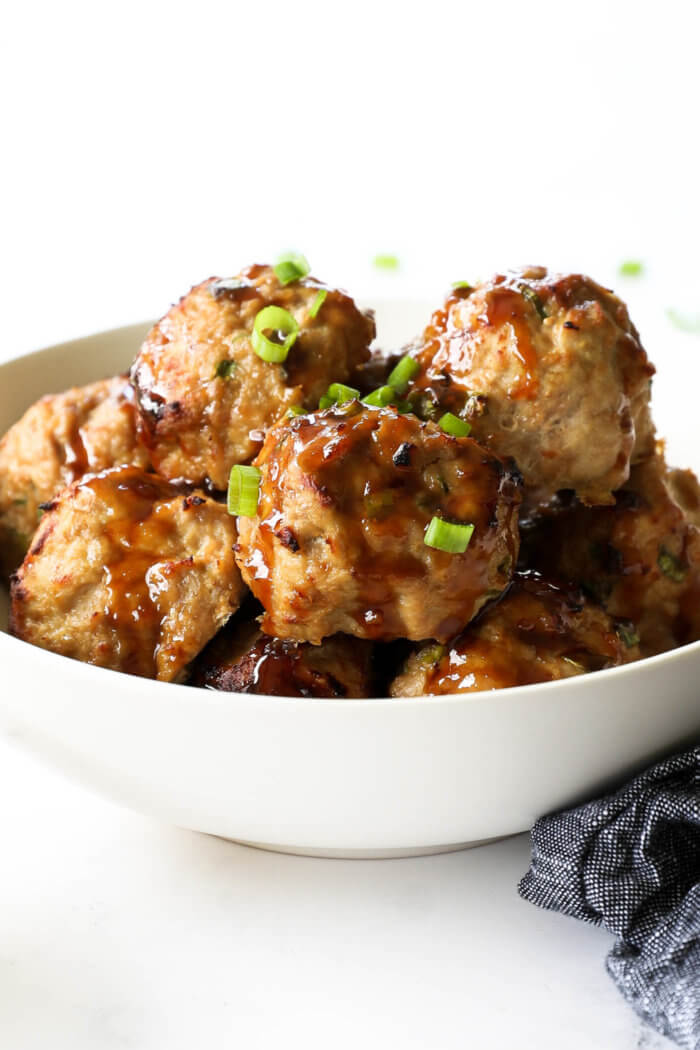 Angled image of a teriyaki turkey meatballs piled in a bowl with sticky teriyaki sauce drizzled on top. Garnished with chopped green onion.