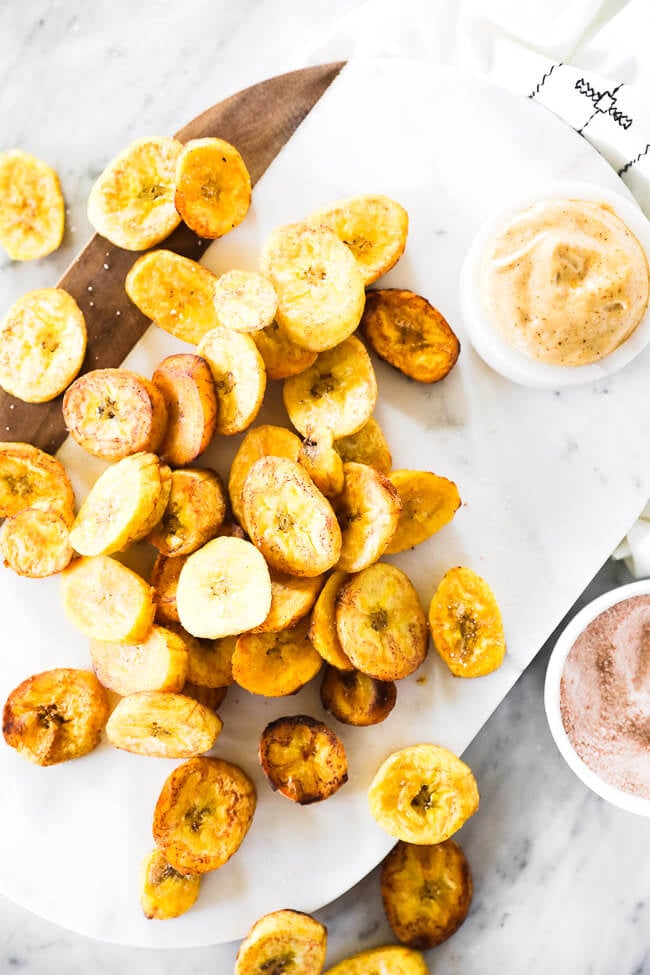 Vertical overhead image of pan fried plantains piled up and scattered on a marble board with dipping sauce and sea salt on the the side. 