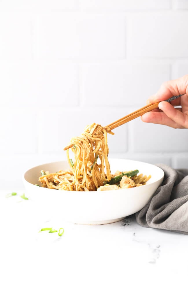 Straight on image of pulling some creamy peanut butter chicken noodles out of a bowl with chopsticks. 