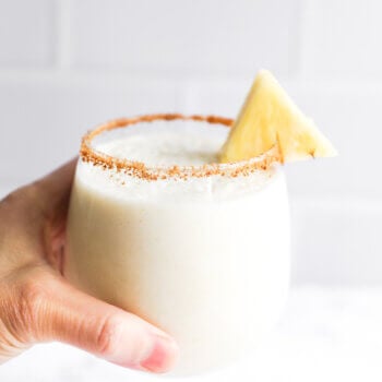Holding a glass with a pina colada mocktail. Glass is lined with coconut sugar and a slice of pineapple.