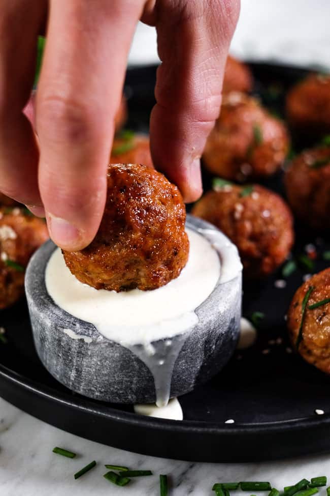 Pork meatball being dipped in ranch dressing with more meatballs in the background. 