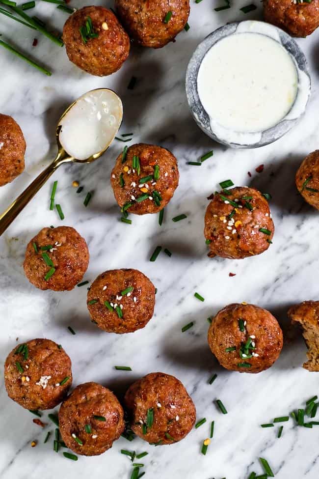 Pork meatballs topped with chopped chives, red pepper chili flakes and sesame seeds spread out on marble with a side of ranch dressing. 