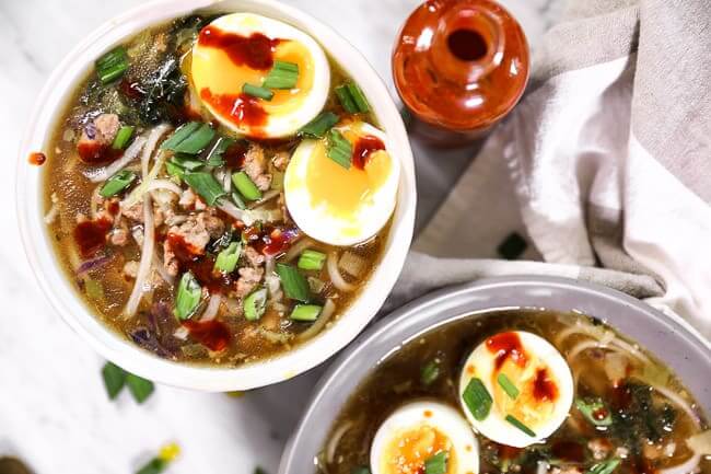 Light and bright bowl of pork ramen noodles topped with chopped green onion, a runny soft boiled egg and a drizzle of hot sauce. Jar of hot sauce on the side. 