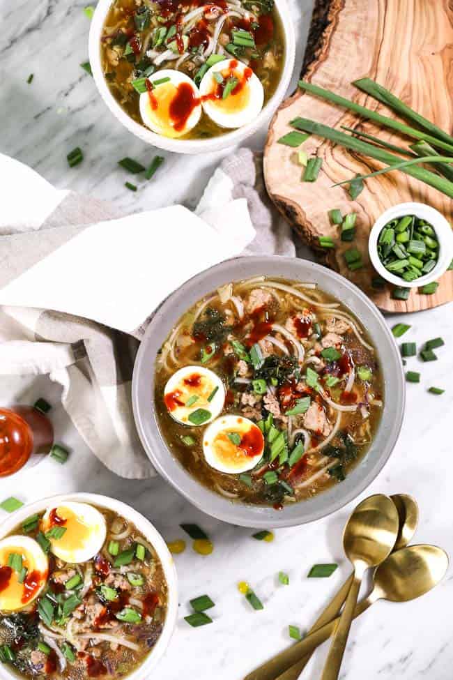 Three bowls of pork ramen noodles, topped with chopped green onion, soft boiled eggs and hot sauce. 