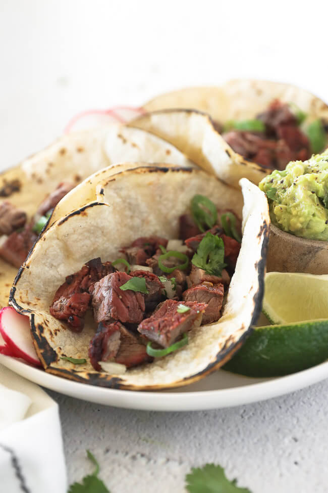 3 carne asada tacos on a plate with cilantro, radish and onion topping