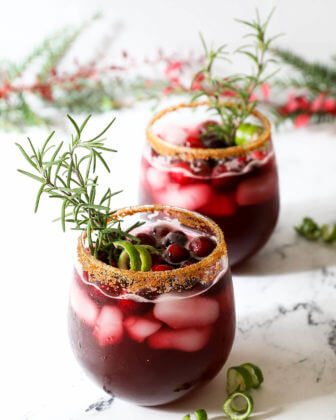 Two glasses of red christmas gin cocktails garnished with rosemary sprigs, lime and cranberries