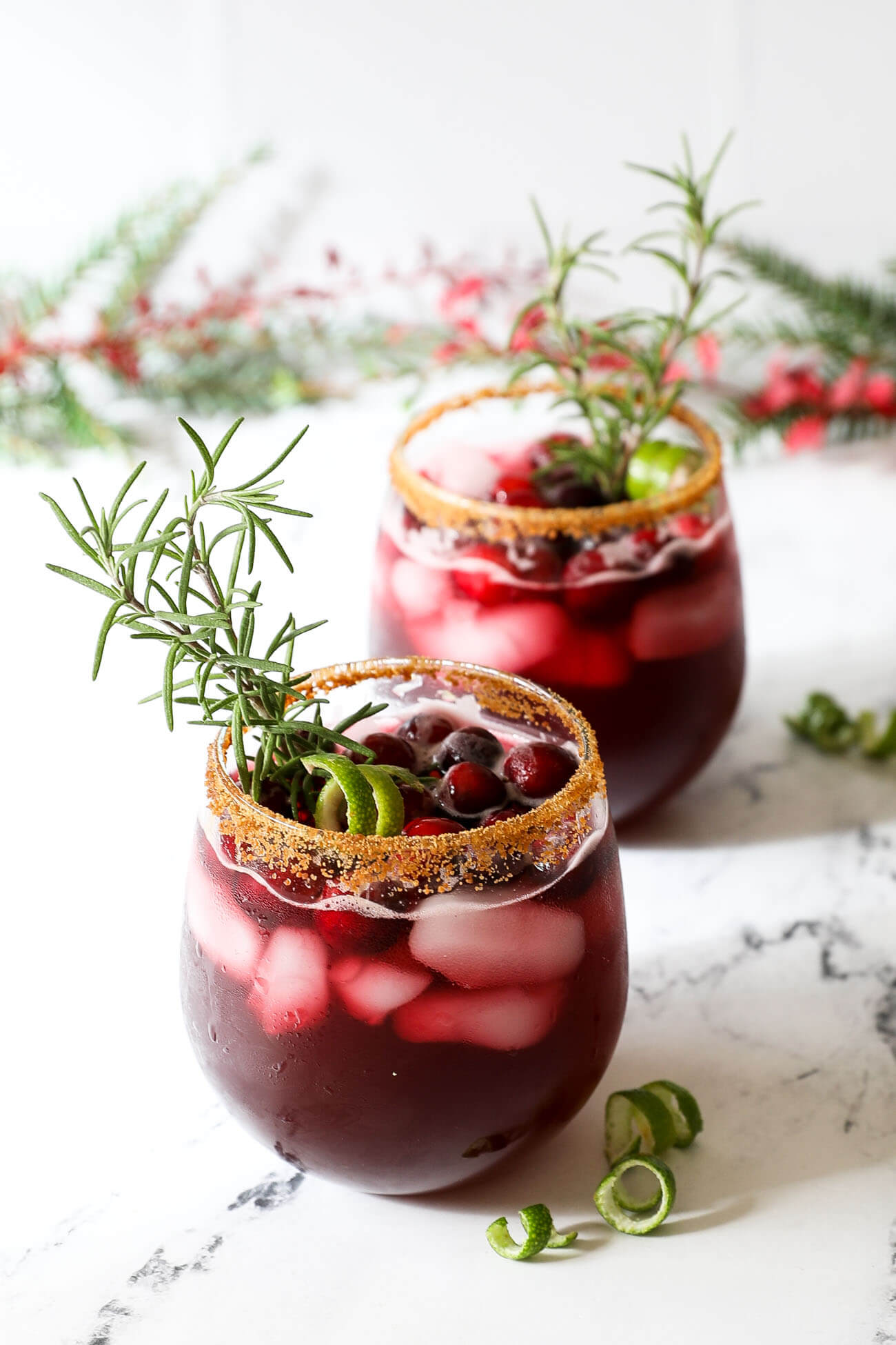 Two glasses of red christmas gin cocktails garnished with rosemary sprigs, lime and cranberries