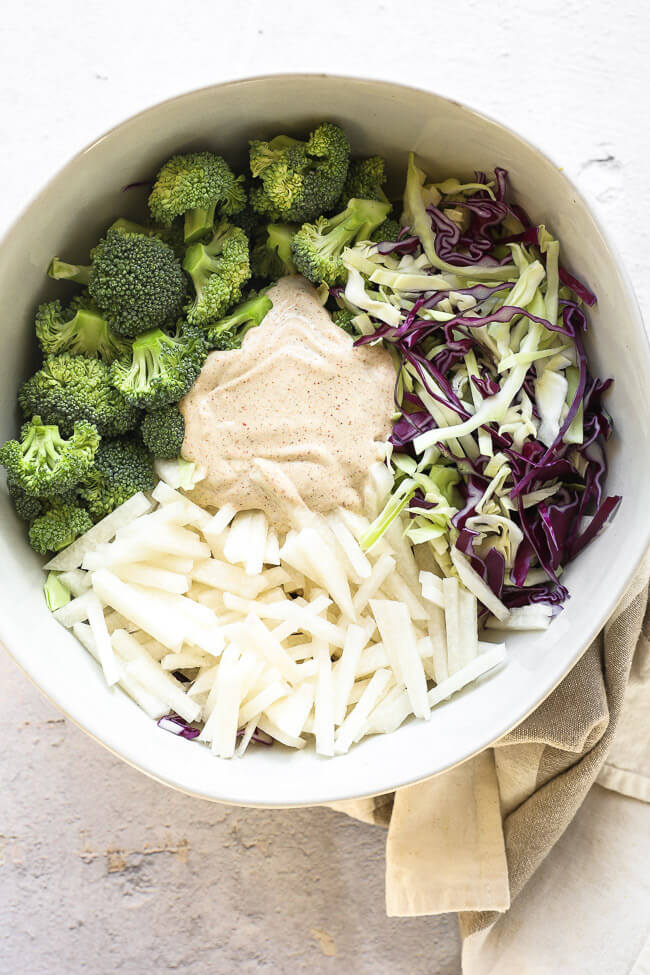 Overhead image of the keto coleslaw ingredients all separated out in a large serving bowl before mixing everything up. Broccoli, purple and green cabbage, jicama and the sauce. 