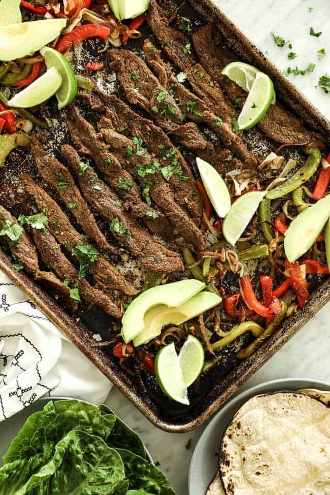 Overhead vertical image of sheet pan fajitas on sheet pan with steak, green and red bell peppers and onion. Topped with fresh cilantro, avocado slices and lime wedges. 