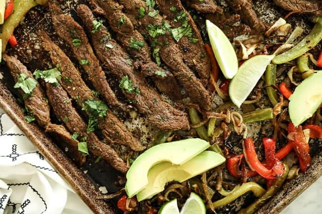 Horizontal overhead image of sheet pan fajitas on sheet pan. Made with steak, green and red bell peppers and onion. Topped with sliced avocados, lime wedges and fresh chopped cilantro. 