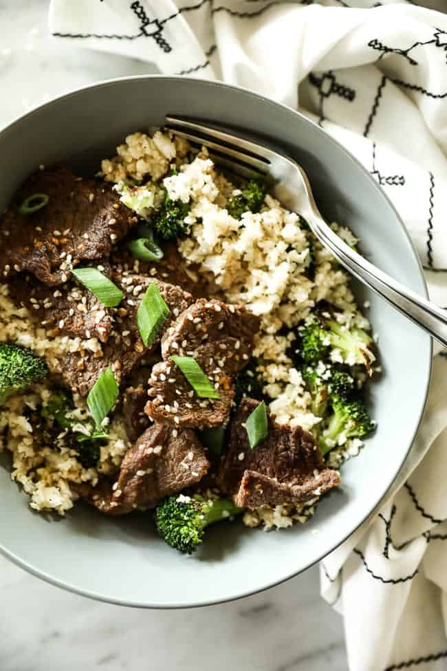 Bowl of cauliflower rice, broccoli and sesame beef with a fork in it. Topped with chopped green onion and sesame seeds. 