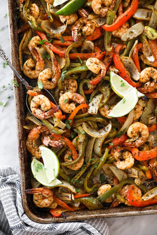 sheet pan shrimp fajitas on a pan with onions, bell pepper and lime wedges overhead vertical image