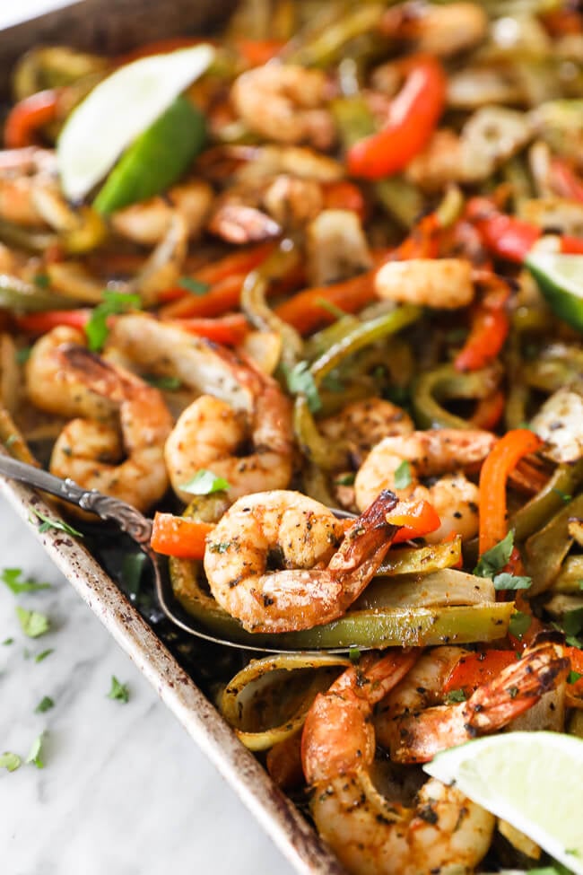 sheet pan shrimp fajitas on a pan with onions, bell pepper and lime. Spoon scooping up shrimp