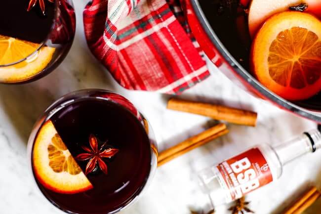 Simple mulled wine recipe in glasses with orange slices, cinnamon sticks, a plaid napkin, star anise and bourbon bottle. 