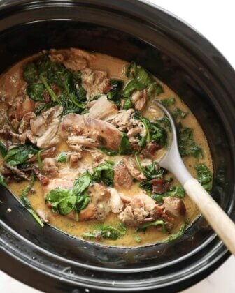 A slow cooker filled with creamy chicken, spinach, and sun-dried tomatoes, with a serving spoon resting inside.