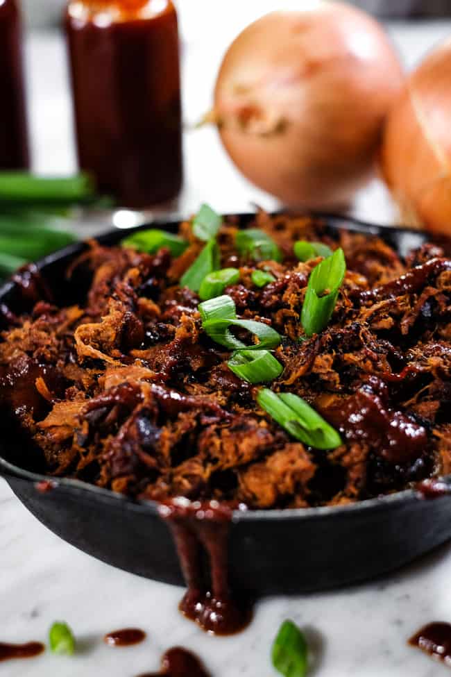 Angle shot of slow cooker BBQ beef in cast iron skillet with extra BBQ sauce and chopped green onion on top. 