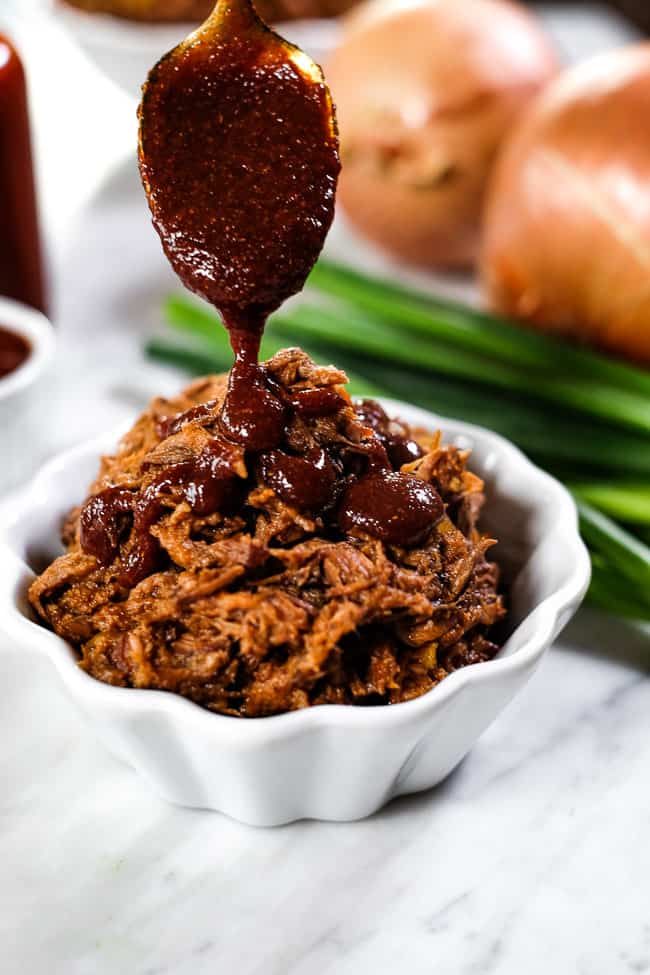 Drizzling BBQ sauce on top of ramekin of slow cooker BBQ beef. Onions in the background. 