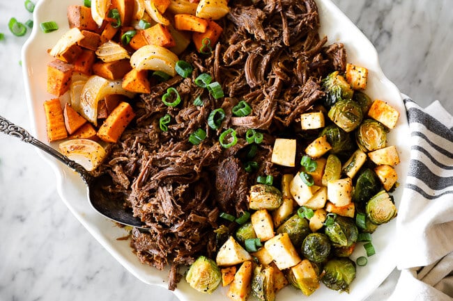 Shredded slow cooker beef pot roast on platter with roasted veggies on each side