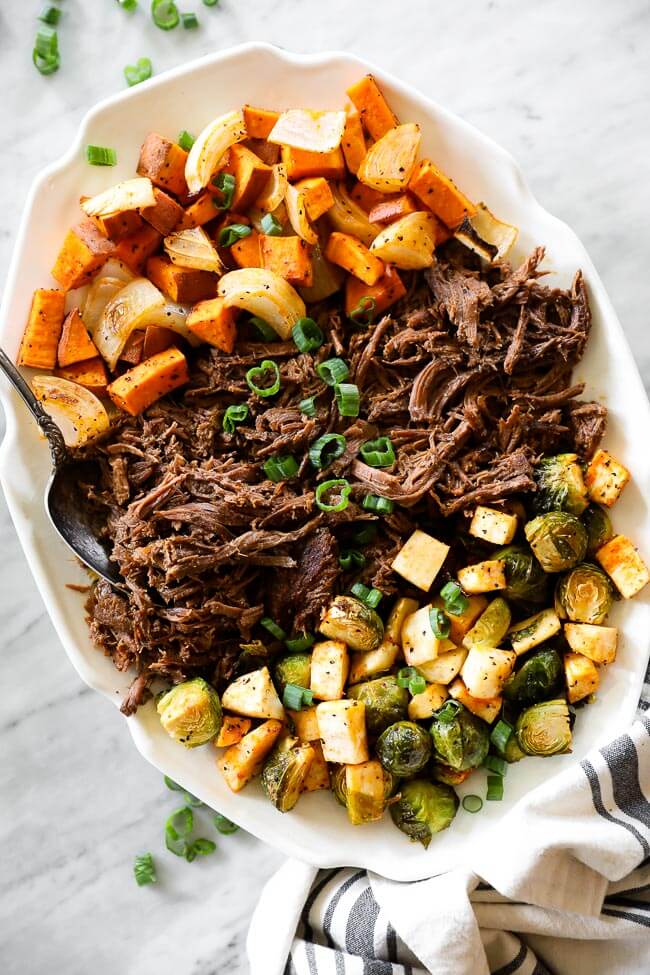 Overhead shot of slow cooker beef pot roast on platter with roasted veggies on each side and spoon on side