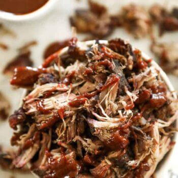 A bowl filled with shredded smoked pork shoulder topped with barbecue sauce, with extra sauce in a bowl nearby.