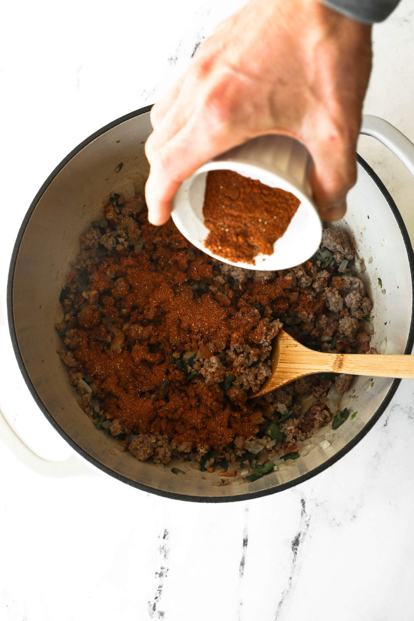 A hand pouring chili seasoning to the ingredients cooking in a dutch oven. 