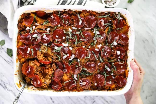 Overhead shot of spaghetti pizza casserole in casserole dish with serving spoon in one corner and holding the dish with one hand. 