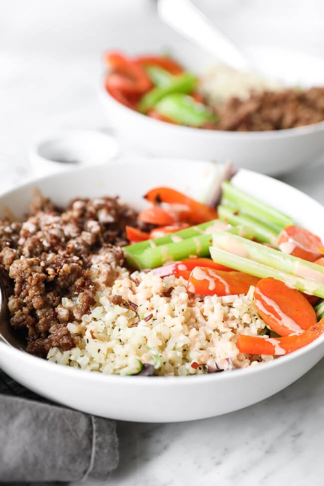 Vertical angled close up shot of bowl filled with spicy pork, cauliflower rice and veggies. Spicy sauce drizzled on top. 