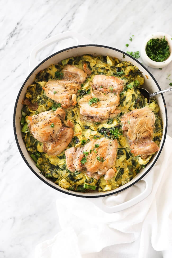 Vertical overhead image of spinach artichoke chicken in a skillet with a serving spoon dug in. Chopped parsley in a small bowl on the side. 