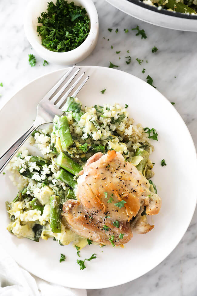 Vertical overhead image of spinach artichoke chicken skillet served on a plate with a fork. Topped with chopped parsley and extra to garnish a small bowl on the side. 