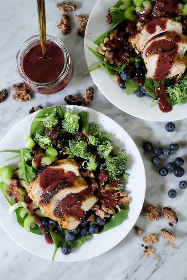 Spinat-blåbærsalat på tallerkener med ekstra valnødder og blåbær spredt rundt omkring og blåbærdressing på siden. Toppet med kylling. 