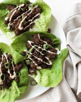 Three lettuce leaves filled with seasoned ground beef, drizzled with a light sauce, and topped with sliced green onions on a white plate next to a beige napkin.