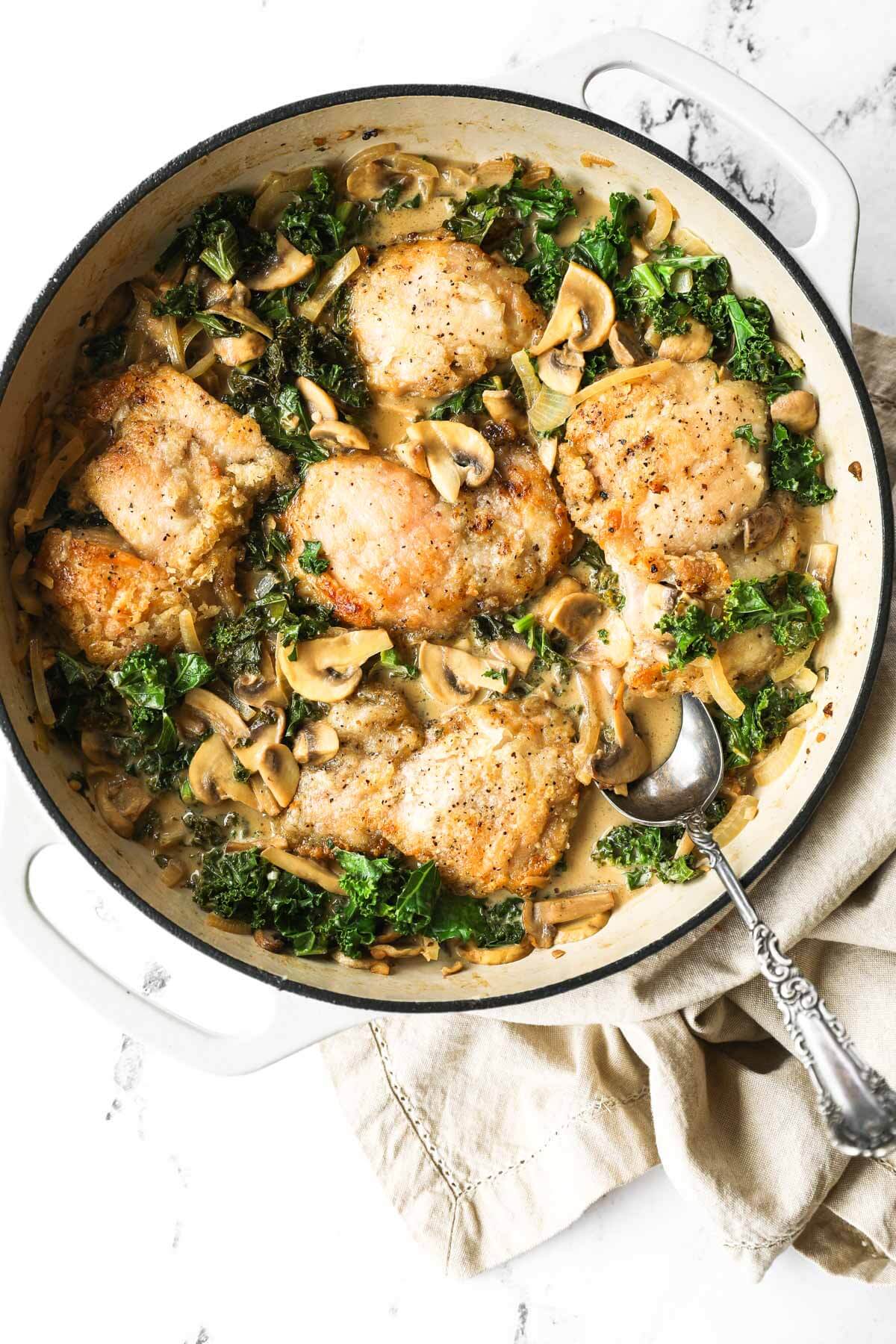 Overhead shot of stovetop creamy mushroom chicken in a pan with kale and mushrooms around chicken and a spoon sticking out