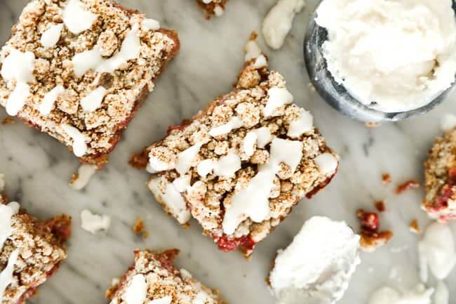 Image of strawberry crumble bars with coconut butter drizzle on top spread out on marble table with a small dish of extra coconut butter and a spoon. 