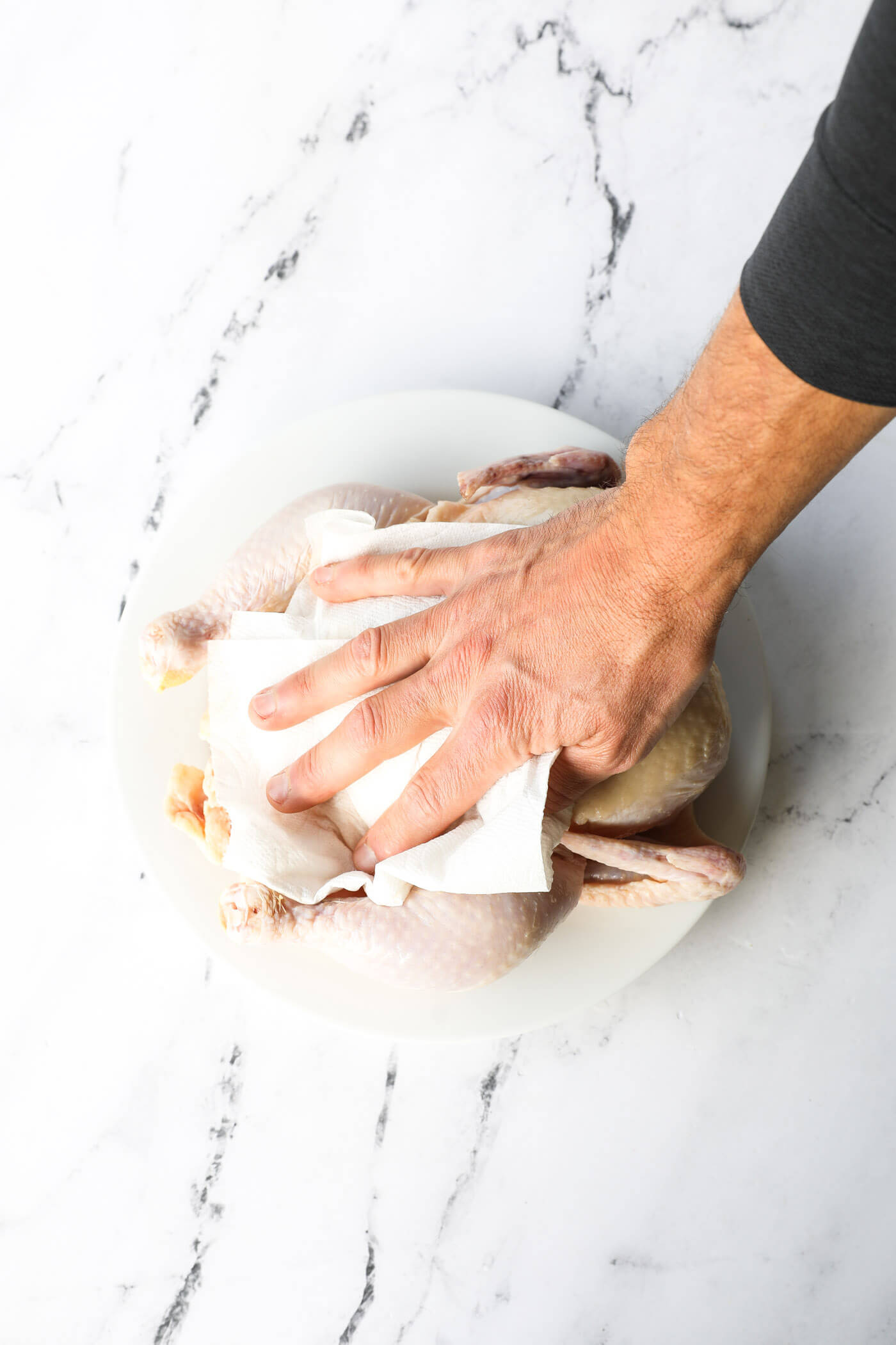 Overhead image of a hand patting a raw whole chicken dry with a paper towel.