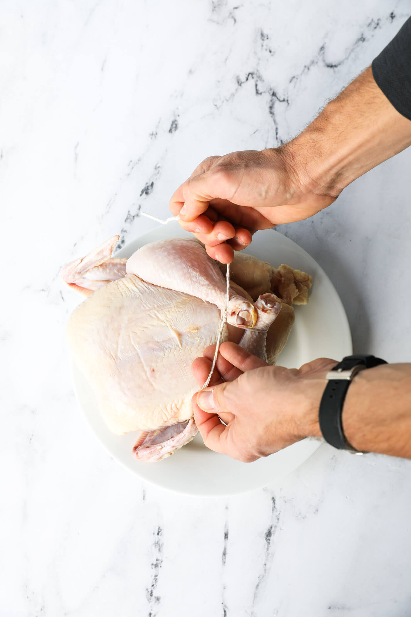 Overhead image of hands tying twine around the legs of a raw whole chicken.