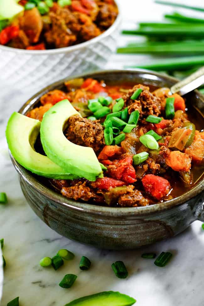 Angled shot of Paleo + Whole30 sweet potato chili in a bowl with chopped green onion and avocado slices. 