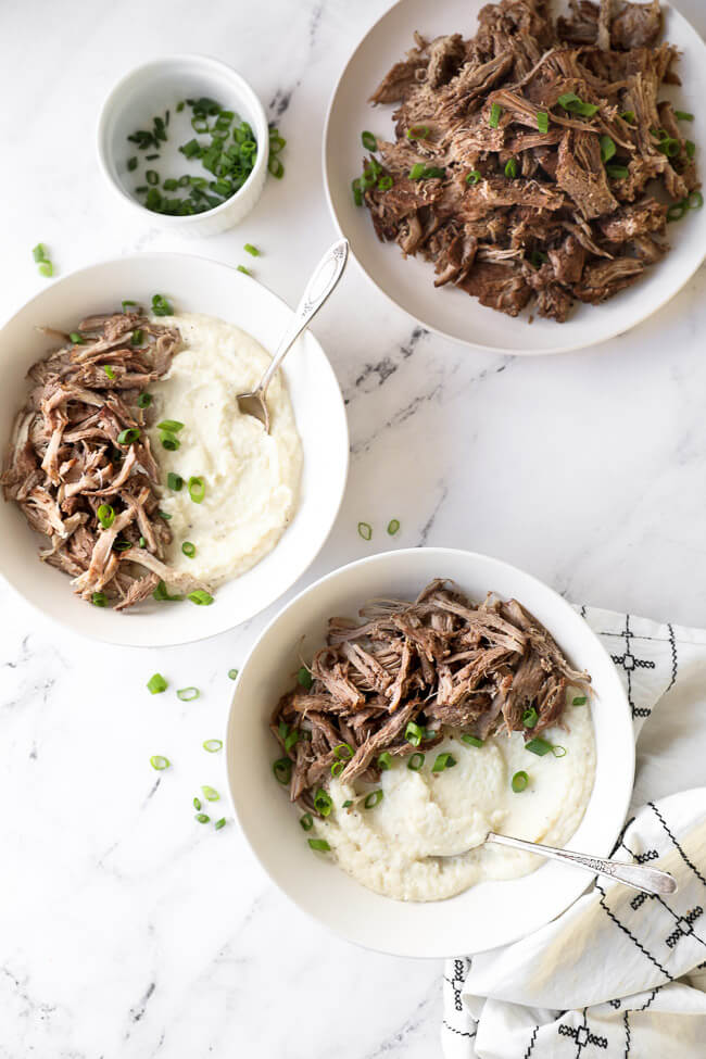 Two bowls of mashed cauliflower and instant pot pork shoulder with green onion on top and forks dug into bowl. Side of extra pulled pork on a plate. 
