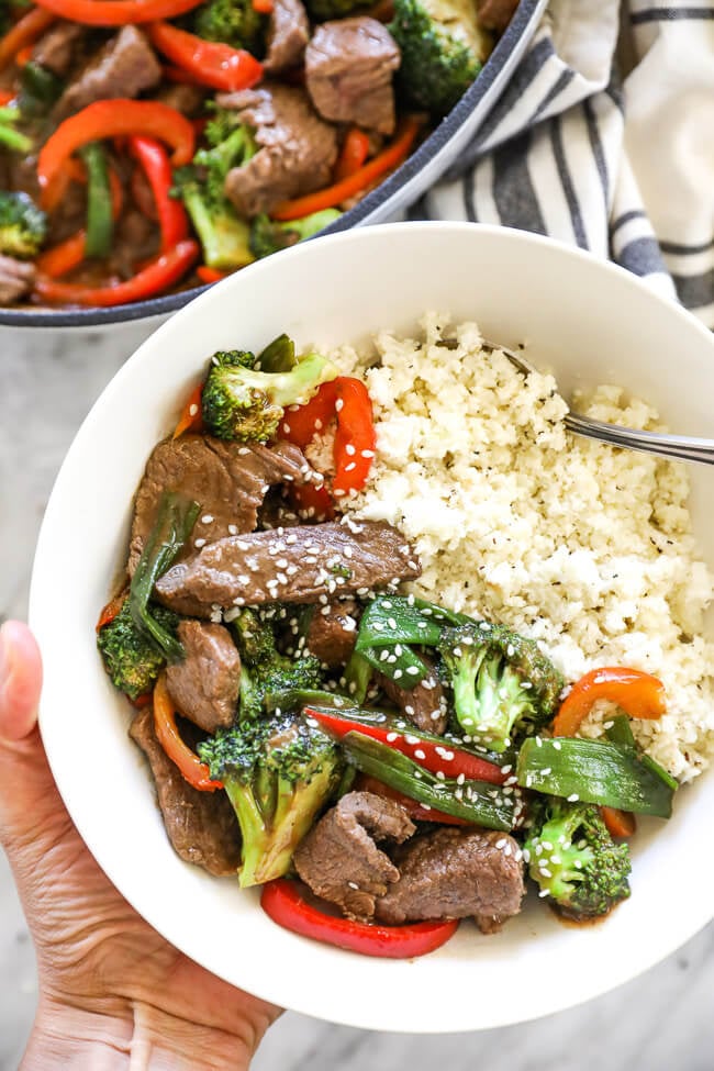 Close up overhead shot of hand holding bowl of teriyaki beef with cauliflower rice and spoon.