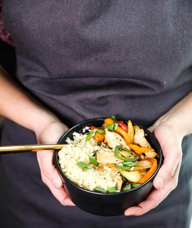 Holding a bowl of Thai basil chicken with a fork in it and topped with chopped green onion. 