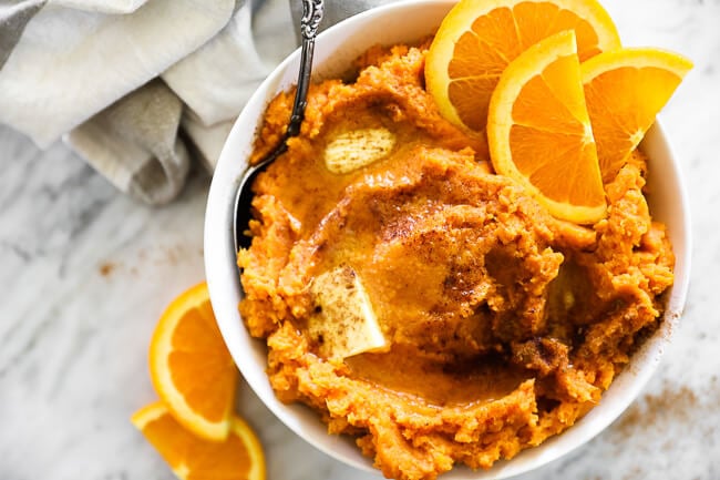 Horizontal overhead image of Thanksgiving yams in a bowl with a serving spoon. Melted butter and cinnamon on top with orange slices as a garnish. 