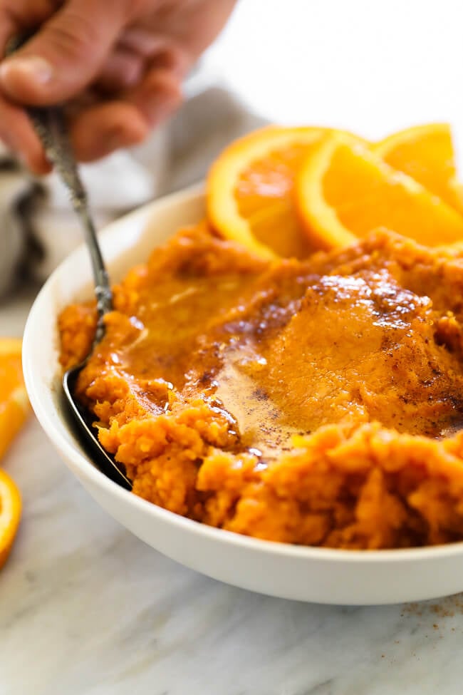 Angled vertical  image of Thanksgiving yams in a bowl with someone taking a scoop out. Melted butter and cinnamon on top. 