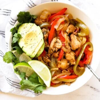 A bowl full of chicken fajitas with sliced avocado, lime wedges and fresh cilantro.