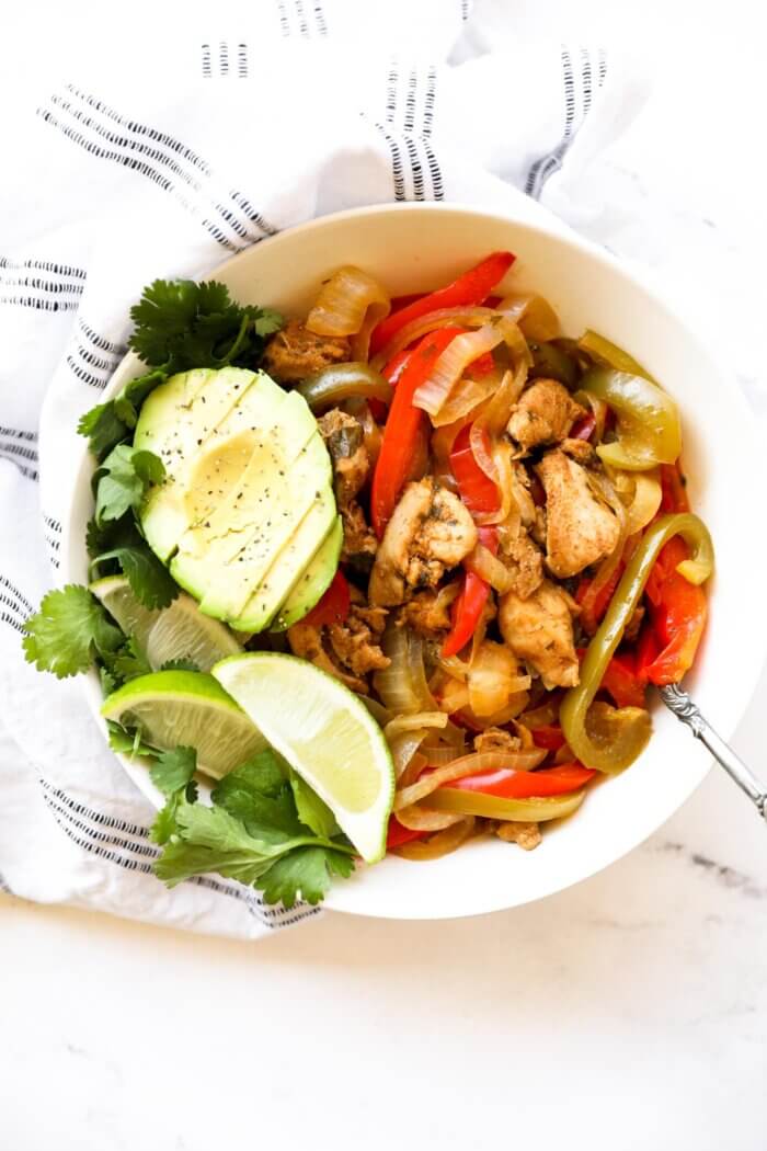 A bowl full of chicken fajitas with sliced avocado, lime wedges and fresh cilantro.