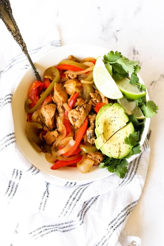 Overhead image of a bowl full of chicken fajitas. Garnished with half an avocado sliced, lime wedges and fresh cilantro. A serving spoon is dug into the bowl.
