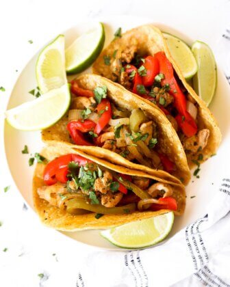 Overhead image of 3 taco shells filled with chicken fajitas. Topped with fresh chopped cilantro and lime wedges holding the tacos up.