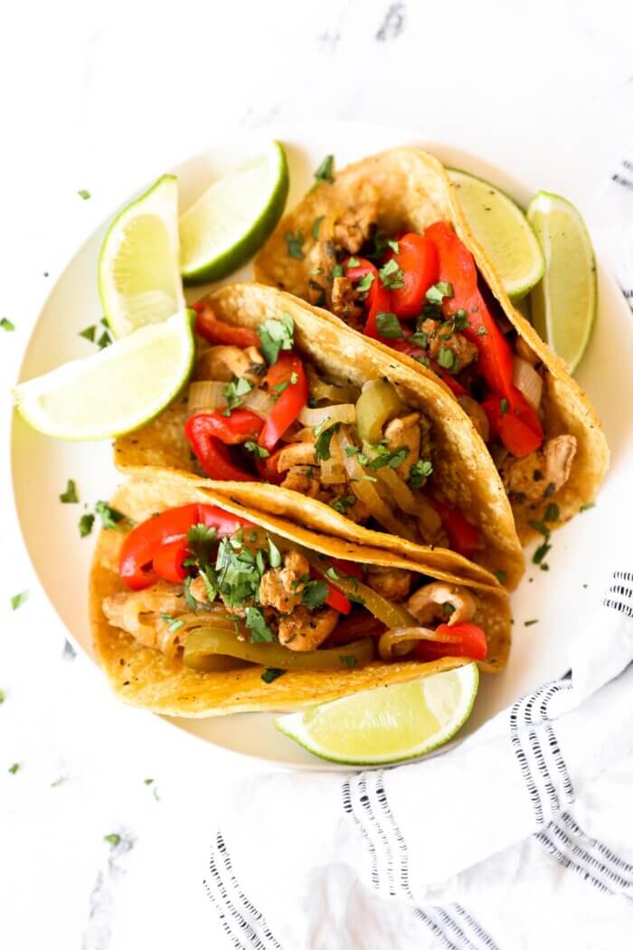 Overhead image of 3 taco shells filled with chicken fajitas. Topped with fresh chopped cilantro and lime wedges holding the tacos up.